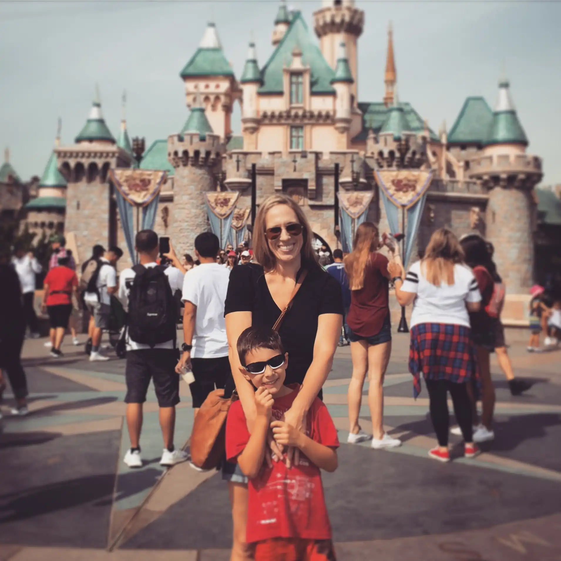 Woman and young boy wearing sunglasses smile while standing in front of a fairytale castle with crowds and banners in the background. The image captures a classic Disneyland memory during a family visit to LaLaLand.