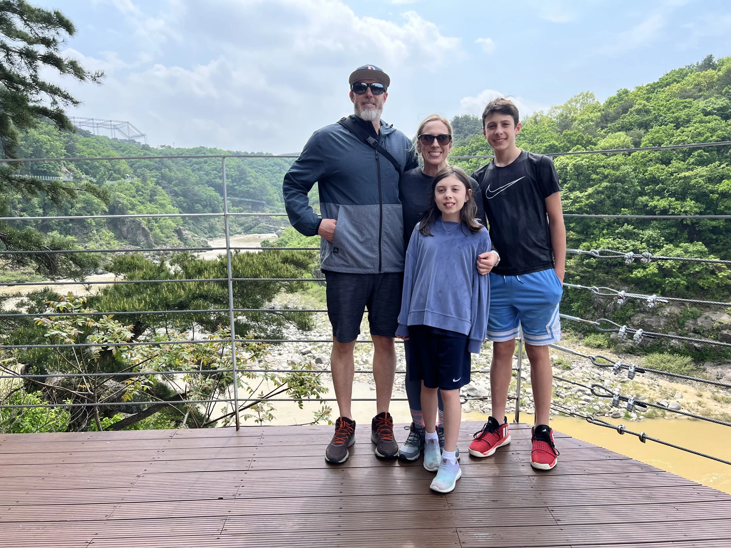 Family of four smiling on a scenic overlook with a river and lush green hills behind them, capturing a moment of togetherness during a move abroad. The group stands on a wooden platform with a suspension bridge visible in the distance, suggesting travel and new beginnings.