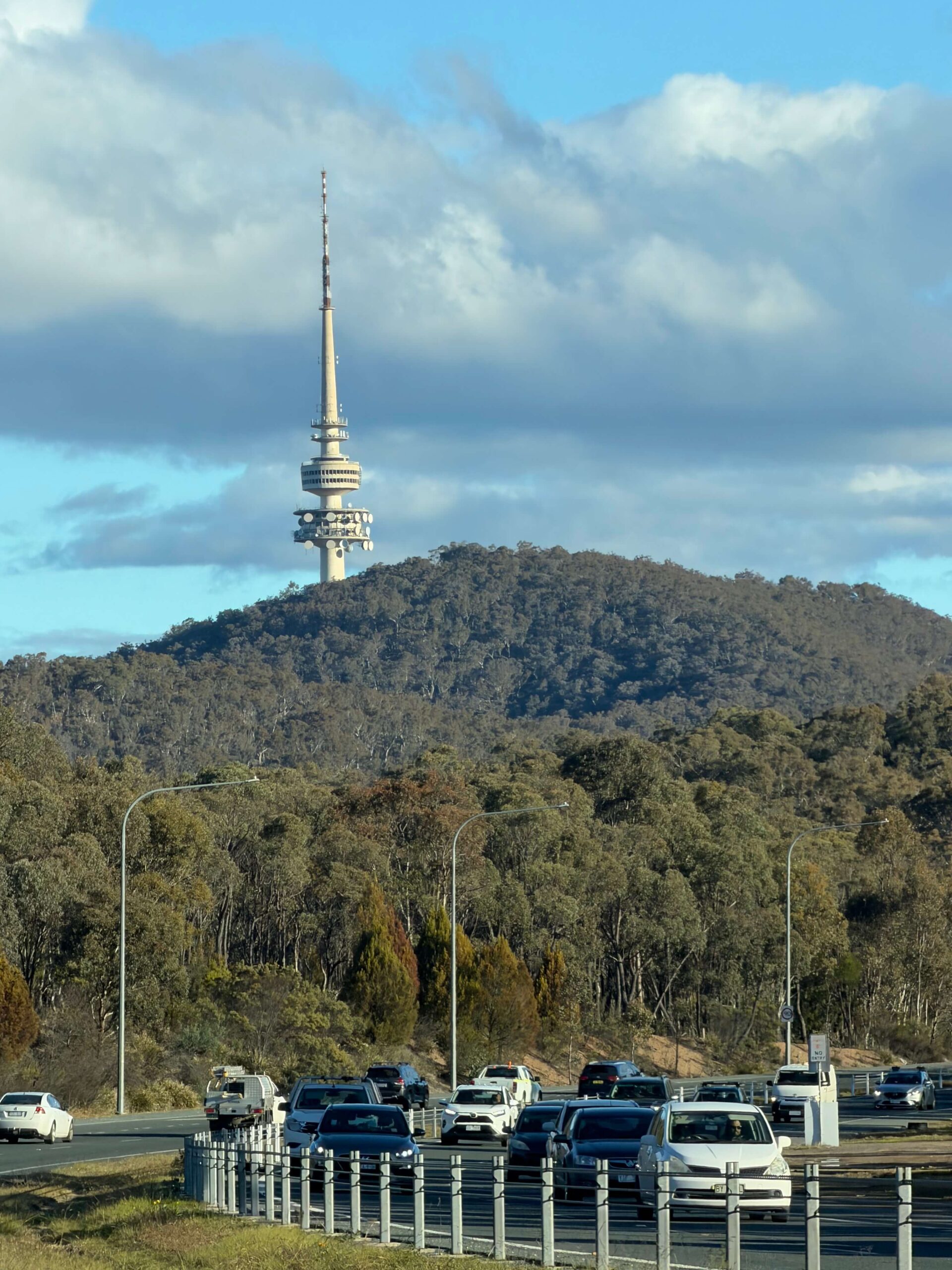 Telstra Tower rises above a densely wooded hill beneath large clouds while traffic travels along the road in the foreground. The landmark is a familiar sight when driving around Canberra.